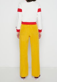 Žluté kordové kalhoty s širokými nohavicemi, zadními kapsami a vertikálním ribbovým vzorem, v kombinaci s bílým topem s červenými pruhy na rukávech.