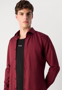 Burgunderfarbendes Hemd mit einer glatten Textur, langen Ärmeln und schwarzen Knöpfen, geschichtet über einem schwarzen Tanktop mit dem Markennamen "HUGO."