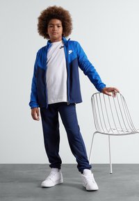 Navy un un zils Nike sporta kostīms ar rāvējslēdzēju jaku un atbilstošām biksēm, zemāk baltā T-kreklā, apvienots ar baltām sporta kurpēm, stāvot blakus baltai krēslam.
