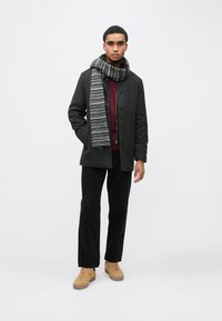 Temno sivi plašč iz volnene mešanice z gumbi spredaj, nošen čez bordo pulover, kombiniran s črnimi hlačami in črno-sivim črtastim šal