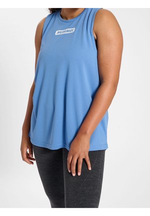 Persona que lleva una camiseta deportiva sin mangas azul de Hummel y leggings texturizados gris oscuro, de pie sobre fondo blanco.