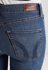 Los jeans de mezclilla azul presentan una etiqueta de cuero marrón con la marca "HOLLISTER", un distintivo cosido en los bolsillos traseros y una silueta ajustada.