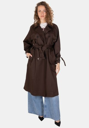 Donna con capelli ricci che indossa un cappotto trench marrone scuro con cintura, jeans a gamba larga azzurri chiari e scarpe nere appuntite, in piedi davanti a uno sfondo bianco.