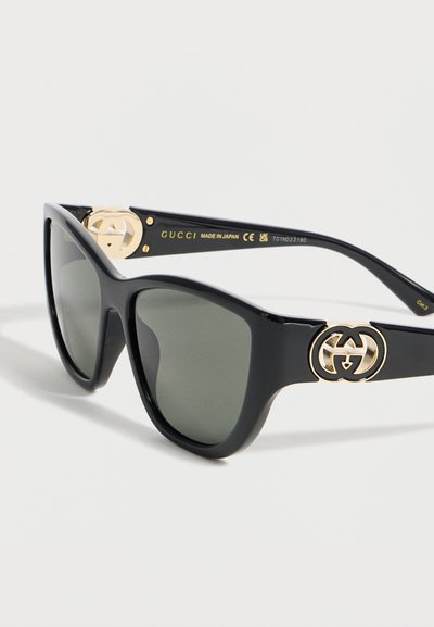 Μαύρα γυαλιά ηλίου cat-eye με σκούρους φακούς και χρυσό λογότυπο Gucci double-G στους βραχίονες, κατασκευασμένα στην Ιαπωνία, ακουμπισμένα σε λευκή επιφάνεια.
