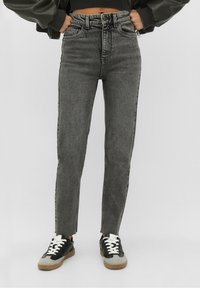 Jeans negros de cintura alta con un ajuste slim, con dobladillo desflecado y rayas laterales en contraste, combinados con zapatillas negras y grises.