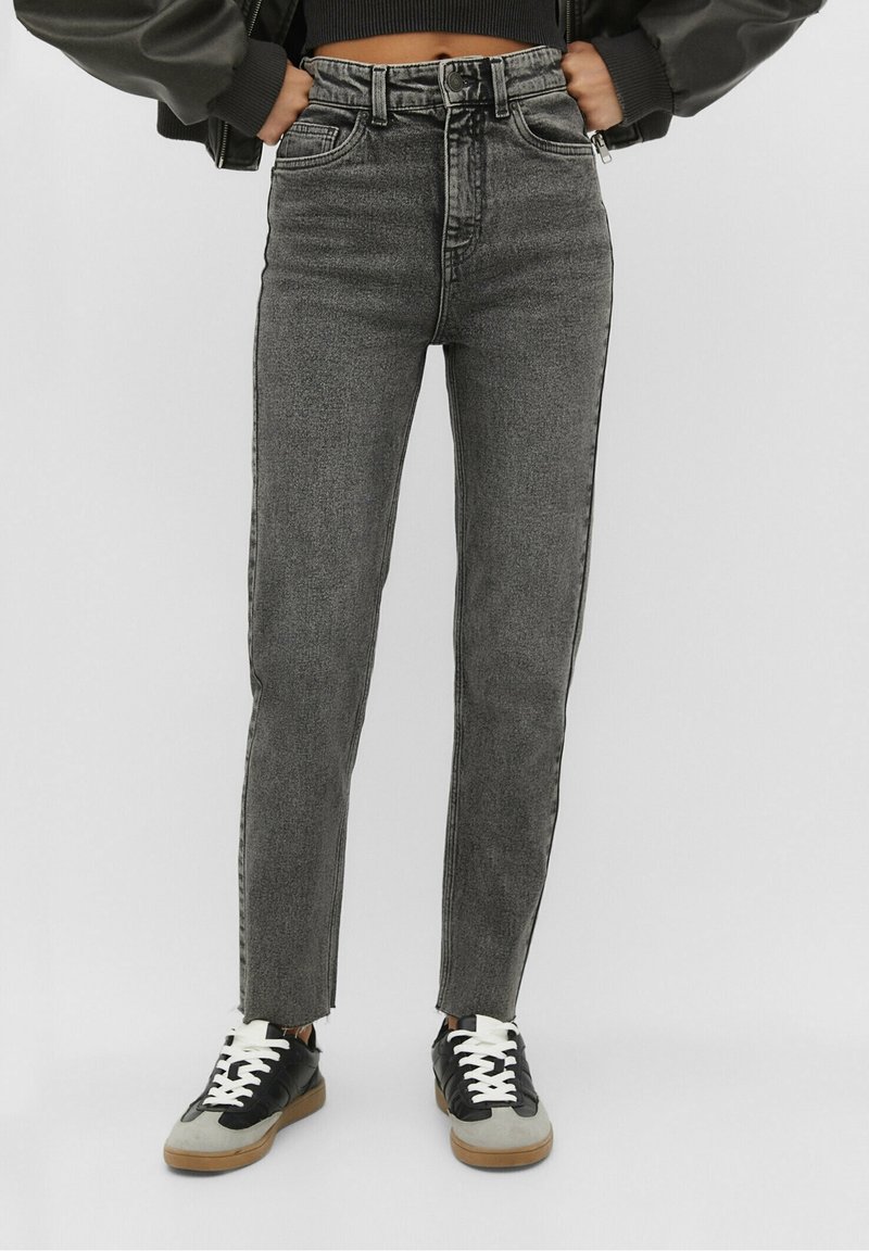 Jeans negros de cintura alta con un ajuste slim, con dobladillo desflecado y rayas laterales en contraste, combinados con zapatillas negras y grises.