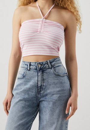 Kobieta ubrana w jasnoróżowy i biało pasiaste halter crop top oraz jasnoniebieskie dżinsy z wysokim stanem na neutralnym tle.