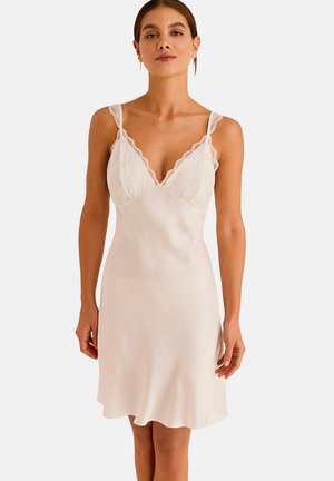 Femme portant une robe nuisette en satin beige clair sans manches avec un bord en dentelle sur le buste et de fines bretelles, debout devant un fond blanc.