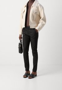 Světle béžová zipová bunda s dvěma předními kapsami, spárovaná s černými tailorovými kalhotami a černými sandály na vysokém podpatku s otevřenou špičkou, držící černou tašku.