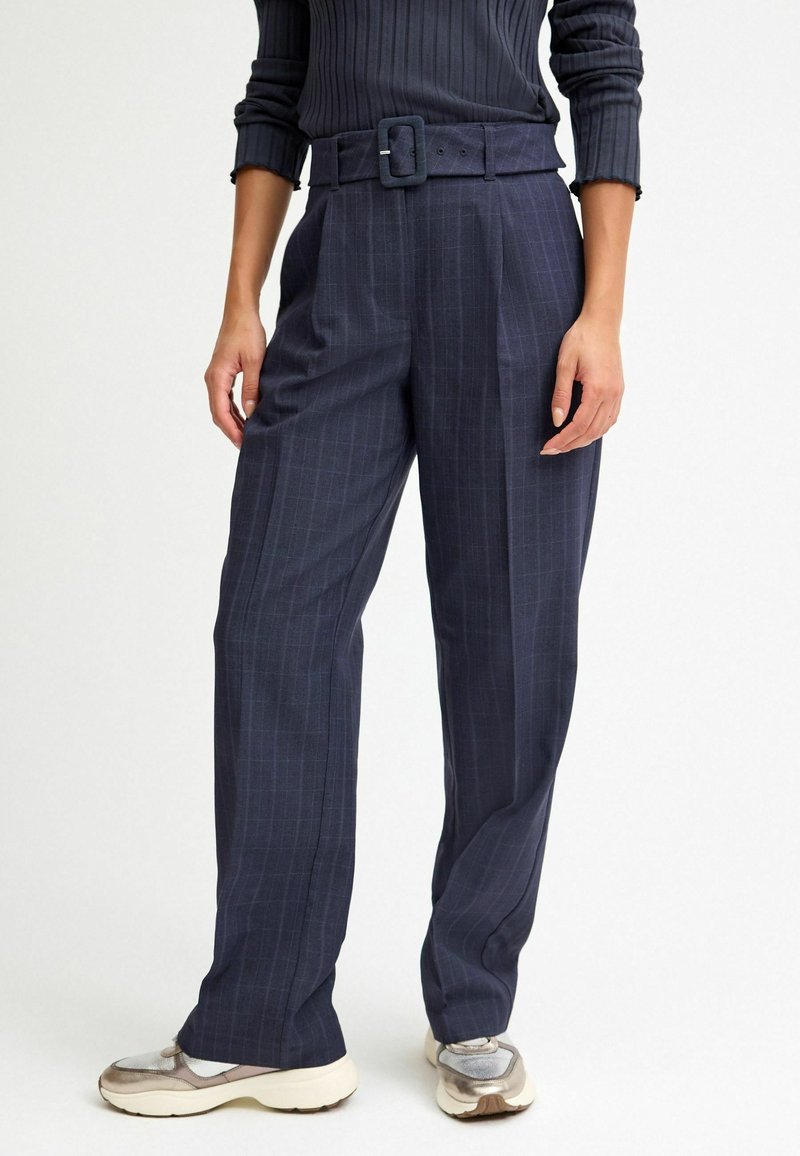 Femme portant un pantalon taille haute bleu marine à fines rayures, avec ceinture et plis, associé à des baskets blanches et grises.