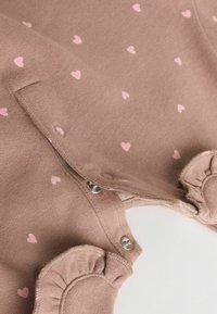 Vêtement en tricot marron avec des motifs de cœurs roses, ailettes froncées aux épaules et pressions sur le côté pour un habillage facile.