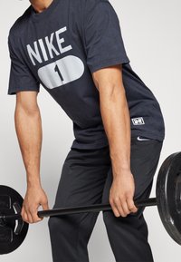 T-shirt Nike bleu marine foncé avec logo blanc sur le devant, associé à un pantalon de sport noir, en soulevant une barre avec des poids ronds.