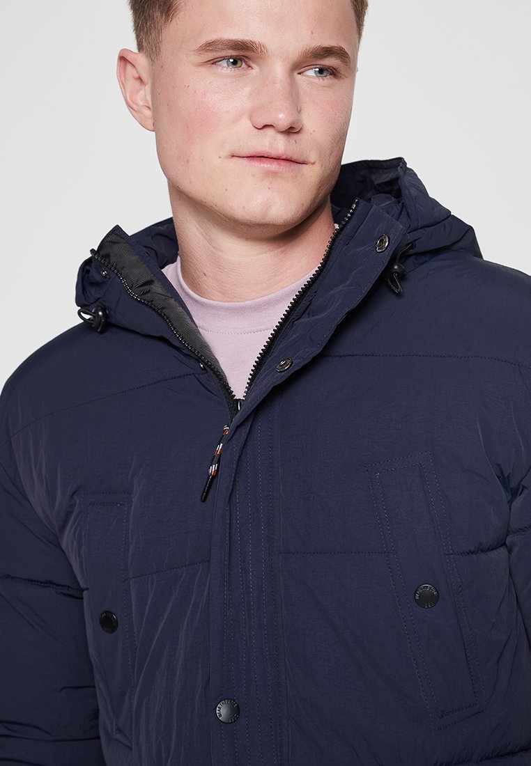 Veste matelassée marine avec capuche, dotée de poches avant, de boutons-pression et d'une fermeture éclair. Texture lisse avec des accents cousus.