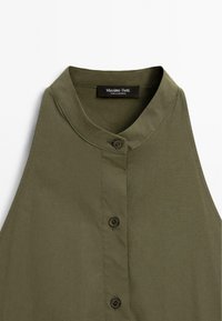 Débardeur vert olive sans manches avec col montant, doté d'une fermeture à boutons et d'une texture de tissu lisse. L'étiquette indique "Massimo Dutti."