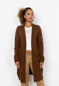Brown long cardigan made of soft knit fabric, featuring ribbed cuffs and pockets, worn over a white shirt and tan pants.
