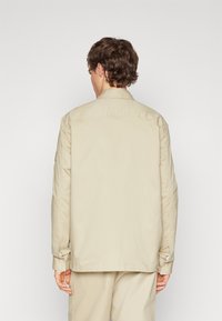 Chaqueta de lona beige con cuello abotonado y mangas largas. Presenta un ajuste holgado y detalles en los puños. Textura lisa, vista trasera.