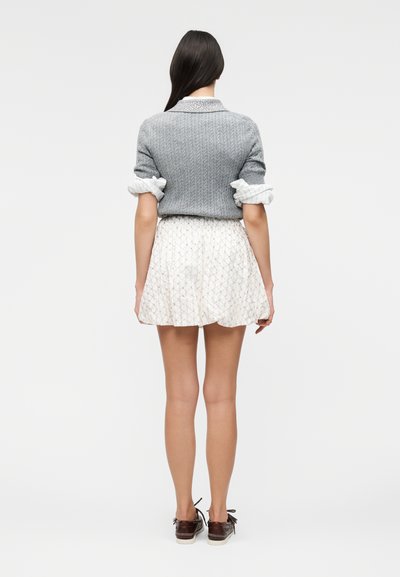 Γυναίκα που στέκεται με την πλάτη προς την κάμερα, φοράει γκρι πλεκτό πουλόβερ με γυρισμένα μανίκια, λευκή φούστα με σχέδια και καφέ παπούτσια με κορδόνια σε μονόχρωμο φόντο.