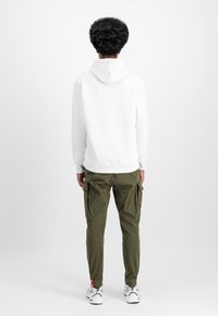 White hoodie with a back pocket and ribbed hem, paired with olive green cargo pants featuring side pockets, elastic cuffs, and red accents.