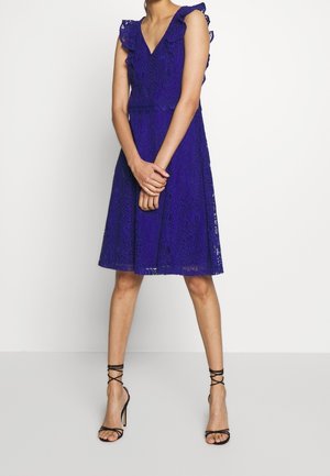 Kobieta w niebieskiej koronkowej sukience do kolan bez rękawów z falbanami na ramionach, nosząca czarne, paskowane szpilki, stoi na białym tle.