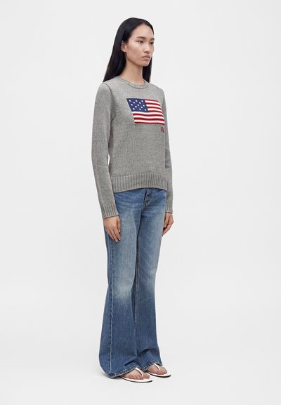 Femme aux longs cheveux foncés portant un pull gris avec un drapeau américain, un jean flare bleu et des sandales blanches, debout devant un fond uni.