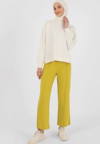 Crème-kleurig sweatshirt met lange mouwen gecombineerd met felgele wijde broek, versterkt met zichtbare stiksels, en witte sneakers.