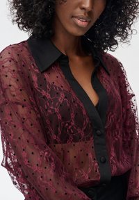 Przezroczysta bordowa bluzka z czarnymi akcentami, zdobiona kwiatową koronką, groszkami, z zapięciem na guziki i klasycznym kołnierzem.