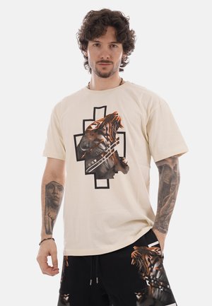 Uomo con capelli ricci e tatuaggi che indossa una maglietta beige e pantaloncini neri, entrambi caratterizzati da grafiche con una tigre ruggente.