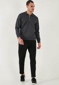 Sötét szürke zipzáros pulóver, puha anyag, hosszú ujjú, fekete slim-fit nadrággal és fekete, tépőzáras sportcipővel párosítva.