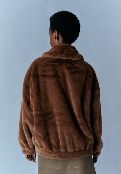 Braune Kunstpelzjacke mit hohem Kragen und elastischem Saum. Glatte Textur und lockere Passform, von hinten vor einem hellen Hintergrund betrachtet.