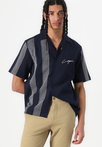 Camisa de manga corta azul marino con rayas verticales blancas y un logo en escritura. Diseño de botones con un corte relajado y textura suave.
