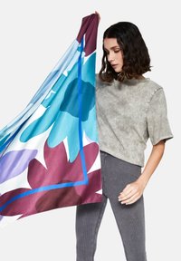 Silk tørklæde med store blomster mønstre i blå, lilla og burgundy, holdt op mod en grå struktureret top og stribede bukser.