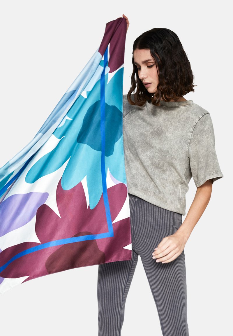 Silk tørklæde med store blomster mønstre i blå, lilla og burgundy, holdt op mod en grå struktureret top og stribede bukser.