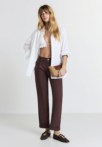 Pantaloni marroni larghi, camicia bianca con bottoni, e un reggiseno bianco arricciato. Tiene una borsa a tracolla intrecciata. Scarpe basse marroni con fori.