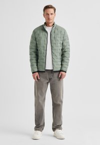 Grüne isolierte Jacke mit gesteppter Optik, Frontreißverschluss und hohem Kragen, kombiniert mit hellgrauen Jeans und weißen Turnschuhen.