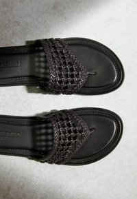 Paire de sandales en cuir marron foncé avec des brides tressées et des boucles pour les orteils, posées à plat sur une surface gris clair.