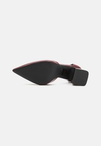 Scarpa con tacco alto in pelle verniciata rossa, con punta affusolata e suola in gomma nera texturizzata, caratterizzata da un design elegante e accenti minimi.