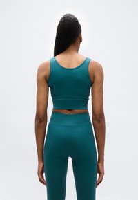 Γυναίκα όρθια, με την πλάτη, φοράει τιρκουάζ ριμπ τοπ χωρίς μανίκια και αντίστοιχο ψηλόμεσο μαύρο κολάν, σε απλό φωτεινό φόντο.