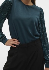 Top turchese con maniche lunghe di paillettes e scollatura rotonda, abbinato a pantaloni neri a vita alta. Texture liscia e vestibilità aderente.