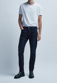 Mörkblå denimjeans med slim fit, kombinerat med en vit t-shirt med rund hals; svarta ankelskot kompletterar outfiten.