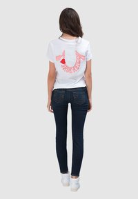 Wit katoenen T-shirt met "True Religion" in het roze gedrukt op de achterkant, gecombineerd met donkerblauwe skinny jeans en witte sneakers.