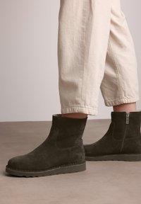 Botas de tobillo de ante negro con suela de goma plana, cremallera lateral y textura suave. Llevadas con pantalones cortos de color claro.