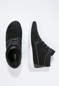 Černé semišové kotníkové boty se šedými tkaničkami. Mají bílou gumovou podrážku, kontrastní švy a měkkou texturu. Logo Ecco viditelné uvnitř.