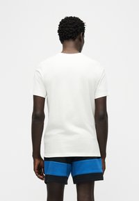 Άνδρας φαίνεται από πίσω φορώντας απλό λευκό κοντομάνικο μπλουζάκι και μαύρο σορτς με φαρδιές μπλε οριζόντιες ρίγες.