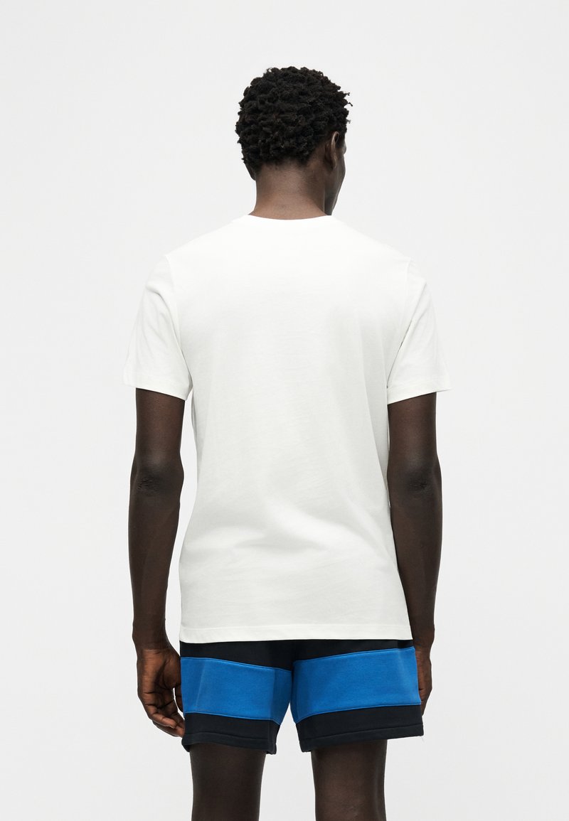 Uomo visto di spalle che indossa una maglietta bianca a maniche corte e pantaloncini neri con ampie strisce orizzontali blu.