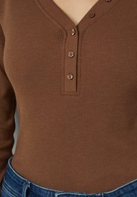 Brązowa prążkowana bluzka z długim rękawem, dekoltem w serek i plisą z pięcioma guzikami, zestawiona z dżinsami, podkreślająca fakturę i dopasowanie.