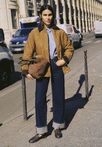 Femme en veste matelassée marron, chemise bleue, jean foncé retourné et chaussures noires, tenant un grand sac marron, debout sur un trottoir en ville près de véhicules stationnés.
