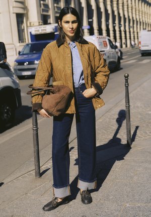 Femme en veste matelassée marron, chemise bleue, jean foncé retourné et chaussures noires, tenant un grand sac marron, debout sur un trottoir en ville près de véhicules stationnés.