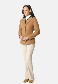 Blazer color camel con capucha negra, dos bolsillos frontales y botones plateados. Lleva puesto sobre un cuello de tortuga blanco, combinado con pantalones beige y botas color canela.