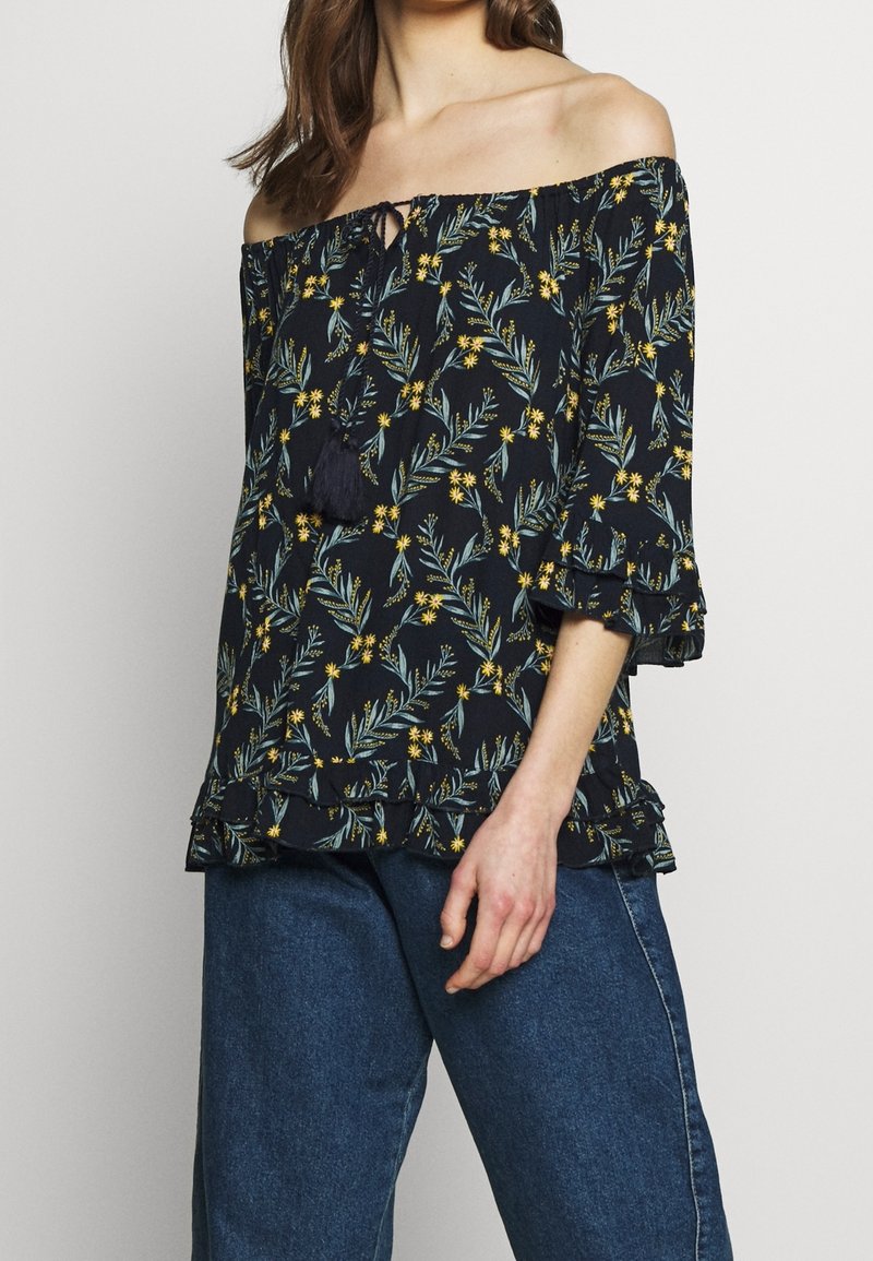 Top off-the-shoulder en azul marino con estampado floral en verdes y amarillos, con detalle de flecos y dobladillo con volantes. Llevado con pantalones de mezclilla azules.