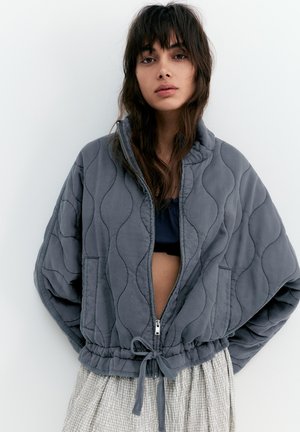 Tânără cu păr ondulat castaniu, purtând o jachetă gri matlasată, desfăcută, peste un top negru și o fustă ușoară în carouri, pe un fundal alb.